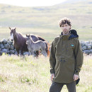 Man standing in a sunlit field wearing a teak brown waterproof smock with a high collar and hood, with horses grazing in the background.