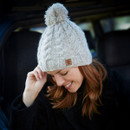 A woman with red hair smiles softly, wearing a cosy beige knit hat with a pom-pom. She is outside, suggesting a chilly environment.