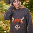 A woman in a cosy, grey knit sweater featuring a cute orange highland cow design, smiles in an autumn park setting. She wears a matching knit hat.
