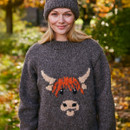 A smiling woman in an autumnal setting wears a gray sweater with a cute orange and beige cow pattern. She's also sporting a matching gray knit hat.