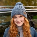 Close-up front view of woman wearing Enniskerry Bobble Beanie in slate blue – Soft hand-knitted wool beanie with pom-pom top and intricate cable knit pattern, styled with a grey turtleneck and denim jacket, ideal for cosy winter wear.