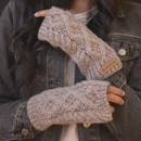 Close-up view of a model’s crossed arms wearing chunky-knit mushroom-coloured fingerless handwarmers with a cable pattern and brown “Pachamama” logo tag, styled with a dark denim jacket.