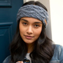 Close-up portrait of a woman wearing a knitted slate blue cable-knit headband by Pachamama, with long dark hair, soft smile, and casual outdoor style.