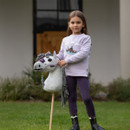A young girl standing on grass holding a grey-and-white spotted hobby horse with a grey yarn mane, wearing a light purple sweatshirt with a horse graphic, purple riding trousers, and black boots, with a blurred stable background.