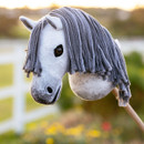 A close-up of a toy hobby horse head with white fabric, black spots, and a long grey yarn mane, mounted on a wooden stick, with a blurred outdoor background featuring a white fence and colorful flowers.