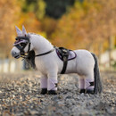 A grey plush toy horse standing on gravel, wearing a full set of miniature tack and accessories in lilac and black, including a crocheted ear bonnet, bridle, quilted saddle pad, saddle, and fleece leg wraps, with an autumnal background of blurred trees.