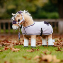 A tan plush toy horse wearing a light purple fleece rug with black trim and a decorative black cord pattern on the hindquarters, a matching purple headcollar, and a purple-and-white lead rope, standing on grass with scattered autumn leaves in the background.