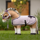 A light brown plush horse toy stands on grass, dressed in a lilac fleece rug with black trim and decorative cord detail on the hindquarters. It wears a matching lilac and purple headcollar with a lead rope attached, and has a black-and-white yarn mane and tail.
