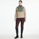 Woman wearing a tan fleece jacket with a sage green quilted front panel, quarter-zip closure, and high collar, paired with burgundy riding tights and black riding boots, standing against a plain white background.