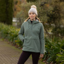 A woman stands outdoors wearing an olive green hooded anorak with a half-zip front, a front flap pocket embroidered with "LeMieux," and black leggings. She has on a light beige knit beanie topped with a large faux fur pom-pom. The background features green foliage and a paved path.
