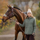 A woman wearing a sage green hooded anorak jacket with a front half-zip and large kangaroo pocket stands outdoors holding the lead rope of a bay horse with a white star marking, maroon halter, and braided mane. She also wears a beige knit beanie with a pom-pom and dark riding tights, with trees and a path in the blurred background.