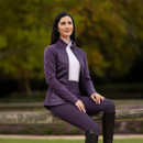 A woman sitting sideways on a wooden fence, wearing a deep purple quilted-front jacket, white base layer, matching riding leggings, and tall black riding boots, with a blurred background of green and autumnal trees. A woman sitting sideways on a wooden fence, wearing a deep purple quilted-front jacket, white base layer, matching riding leggings, and tall black riding boots, with a blurred background of green and autumnal trees.