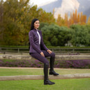 A wider shot of the woman in the purple quilted-front jacket and matching riding leggings, sitting on a wooden fence in a grassy paddock, with green trees and purple flowers in the background. A wider shot of the woman in the purple quilted-front jacket and matching riding leggings, sitting on a wooden fence in a grassy paddock, with green trees and purple flowers in the background.