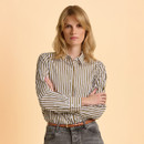 A woman with blonde hair stands against a light peach background, wearing a white and olive striped shirt, grey jeans, and a leather belt. She has an assertive expression with arms crossed.