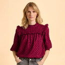 A woman with wavy blonde hair wears a vibrant maroon blouse with textured patterns and ruffle details. She stands confidently against a pale peach background.