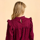 A woman with long blonde hair is facing away, wearing a textured burgundy blouse with ruffled shoulders against a soft beige background.