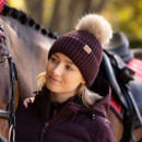 Woman wearing a damson ribbed knit beanie with beige faux fur pom-pom, paired with a matching jacket, standing beside a saddled horse.