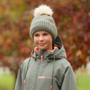 Young woman outdoors in light rain wearing a khaki green ribbed knit beanie with beige faux fur pom pom and matching waterproof jacket with coral accents.