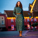 A woman in a green patterned dress and brown boots stands smiling on a street at dusk. Warm lights adorn buildings in the background, creating a cosy, festive atmosphere.