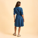 A woman in a denim shirt dress stands facing away on a light beige background, showcasing relaxed elegance with casual trainers and rolled-up sleeves.