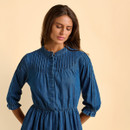 A woman in a blue denim dress and beige sneakers stands against a light peach background, looking downward thoughtfully with a relaxed posture.