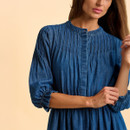 A woman in a denim dress with pleats and ruched sleeves poses against a soft beige background. The tone is casual and stylish.