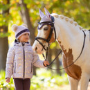 Young girl in lilac LeMieux padded jacket with horse print, wearing purple riding hat and leading a plaited pony in matching lilac ear bonnet.