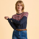 A person wearing a navy cardigan with a colourful patterned yoke, tucked into blue jeans with a brown braided belt. The backdrop is a soft beige, creating a calm tone.