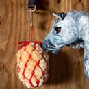 Grey plush toy pony interacting with cream plush haynet ball in red rope net, hanging from a stable wall.