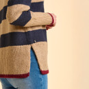 Close-up of a person in a beige and navy striped sweater with red trim, paired with blue jeans. The sweater has a slit on the side, creating a casual look.