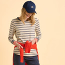 A woman in a navy cap and striped shirt smiles while tying a bright orange jumper around her waist. The background is a warm beige, creating a cheerful tone.