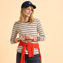 A woman in a navy baseball cap and striped long-sleeve shirt stands against a beige background. A red jumper is tied around her waist, conveying a casual, relaxed vibe.