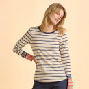 A young woman, wearing a cream and navy striped long-sleeve shirt with contrasting red and navy cuffs, smiles softly against a beige background.