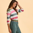 Woman smiling in a pastel-striped shirt and teal pants, hand in hair, set against a soft peach background, conveying a cheerful, relaxed mood.