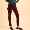 A person wearing maroon corduroy pants stands with hands on hips. They're paired with a blue patterned blouse and brown loafers on a beige background.