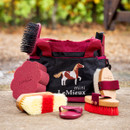 Horse grooming kit with red brushes, sponge, and curry comb in a black and red bag featuring a horse illustration, set against a blurred outdoor background.