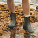Person wearing short chocolate brown Joules Oakfield neoprene wellington boots while standing on wet rocks by the water.