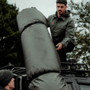 A man in a green jacket is adjusting a large, cylindrical black bag on a vehicle roof rack. The mood is focused, with trees in the background.