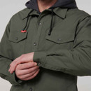 A person wearing an olive green button-up shirt with a black hooded sweatshirt underneath adjusts the cuff. The shirt has buttoned chest pockets.