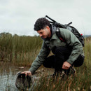 A man in outdoor gear crouches by a pond, inspecting a metal trap in tall grass. He looks focused and the sky is overcast, suggesting a calm, explorative mood.