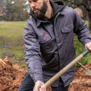 A person in a gray jacket works intently, holding a shovel over a mulch pile. The background features green grass and trees, conveying a focused and diligent mood.