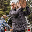 A man with a beard carries a large log on his shoulder with a focused expression. Another person in a plaid jacket is unloading a truck in a forest.