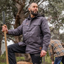 A bearded man stands confidently outdoors, holding a shovel, wearing a gray jacket. Trees and another person in a plaid shirt are visible in the background.