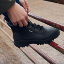 A person tightening the laces on a pair of Barbour Marlow black lace-up wellington boots, designed for waterproof outdoor wear.