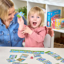 A joyful child, holding a colourful card, sits with an adult at a table. They are playing a board game, surrounded by cards and a dice, in a playful setting.