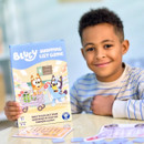 A smiling child in a striped shirt holds a "Bluey Shopping List Game" box featuring colourful characters. The scene conveys fun and excitement.