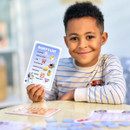 A smiling child in a striped shirt holds up a card titled "Bluey's List," featuring pictures of food and toys. They sit at a table with game pieces.