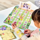 Child playing a colourful board game with image cards on a table. A game board shows various objects; a tablet displays a cartoon. Fun, engaging setting.