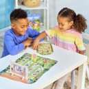 Two children, smiling and engaged, play a board game at a white table. The game has colourful pieces and a map. The atmosphere is joyful and playful.
