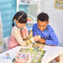 Two children, a girl and a boy, happily play a board game at a table. The setting is colourful and cheerful, with toys and a shelf in the background.